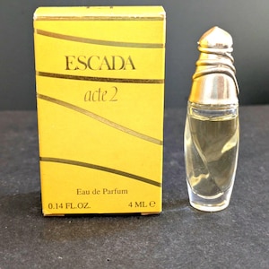 Puede incluir: Un conjunto de perfume en miniatura y su caja. La caja es amarilla con detalles dorados y el texto "ESCADA acte 2". El frasco de perfume es transparente con una tapa plateada y contiene un líquido claro. La caja indica Eau de Parfum, 4 ml.