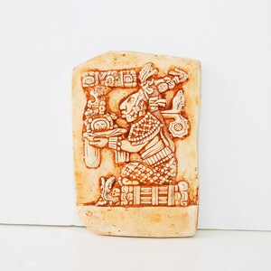Può includere: Una scultura in ceramica beige a rilievo di una figura Maya che tiene una ciotola. La figura indossa un copricapo e un abito a motivi. La scultura è decorata con dettagli intricati e ha una patina marrone.