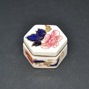 Puede incluir: Una caja de porcelana hexagonal con un diseño floral. La tapa presenta una flor rosa y azul con detalles dorados. La caja está decorada con una cinta azul y dorada.
