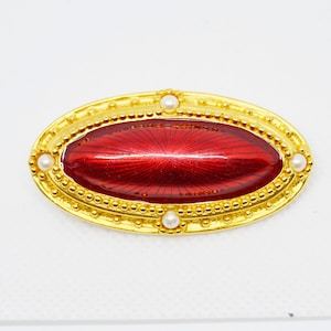 May include: A gold oval brooch with a red enamel center and white pearl accents.