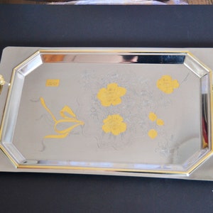 May include: A silver serving tray with gold trim and a floral design. The tray has two gold handles with red accents.