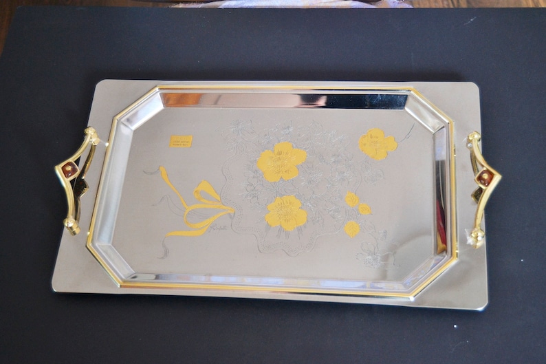 May include: A silver serving tray with gold trim and a floral design. The tray has two gold handles with red accents.
