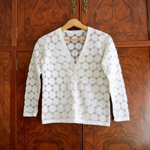 May include: White crochet cardigan with a V-neck and button closure. The cardigan is made of a delicate, open-stitch pattern.