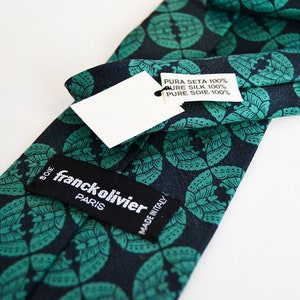 May include: A dark blue silk tie with a green floral pattern. The tie has a label that reads "franck olivier PARIS" and another label that reads "PURA SETA 100% PURE SILK 100% PURE SOIE 100%" and "MADE IN ITALY".