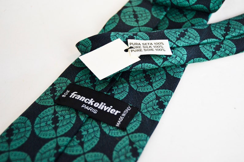 May include: A dark blue silk tie with a green floral pattern. The tie has a label that reads "franck olivier PARIS" and another label that reads "PURA SETA 100% PURE SILK 100% PURE SOIE 100%" and "MADE IN ITALY".