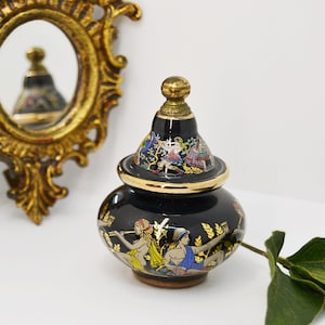 May include: A black and gold ceramic bottle with a lid and a painted design of people and flowers. The bottle is sitting on a white surface.