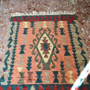 May include: A colourful rug with a geometric pattern in shades of red, green, orange, and brown. The rug has a fringed border and is made of woven wool.