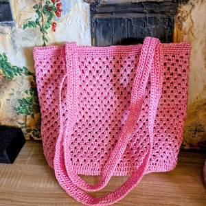 Op de afbeelding: Een roze gehaakte tote bag met lange hengsels. De tas heeft een open geweven ontwerp en is gemaakt van een zacht, getextureerd garen. De tas staat op een houten oppervlak.