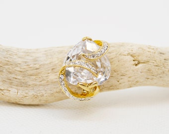 Op de afbeelding: Een gouden ring met een grote, heldere, peervormige steen die in een swirl-ontwerp is gezet. De ring ligt op een lichtbruin houten oppervlak.