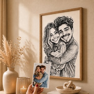 May include: A framed black and white charcoal-style portrait illustration of a smiling couple embracing, displayed on a wall. A smaller colour photograph of the same couple is held in the foreground, showing them in a similar pose wearing denim.