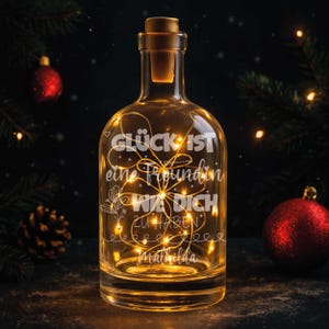 Puede incluir: Una botella de vidrio transparente con tapón de corcho, iluminada por luces de hadas blancas cálidas. La botella tiene texto grabado en alemán. El fondo incluye adornos navideños y vegetación, creando un ambiente festivo.