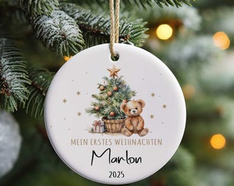 Könnte beinhalten: Weißer Weihnachtsbaumschmuck aus Keramik mit einer Aquarellillustration eines Weihnachtsbaums, eines Teddybären und Geschenken. Der Text lautet "Mein erstes Weihnachten" und "Marlon 2025". Der Schmuck hängt an einer natürlichen Schnur.