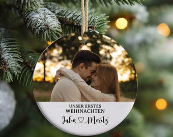 Könnte beinhalten: Runder Weihnachtsschmuck mit einem Foto eines sich umarmenden Paares vor verschwommenem Hintergrund. Der Schmuck trägt den Text "UNSER ERSTES WEIHNACHTEN" und die Namen "Julia & Moritz" in Schriftform. Er hängt an einer Schnur.