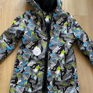 Peut inclure: Une veste à capuche grise avec une doublure en polaire noire et une fermeture éclair frontale. L'extérieur présente un motif répétitif de silhouettes noires de snowboarders, des accents bleus et verts, et le texte EXTREME, SPORT STYLE, LET'S GO.