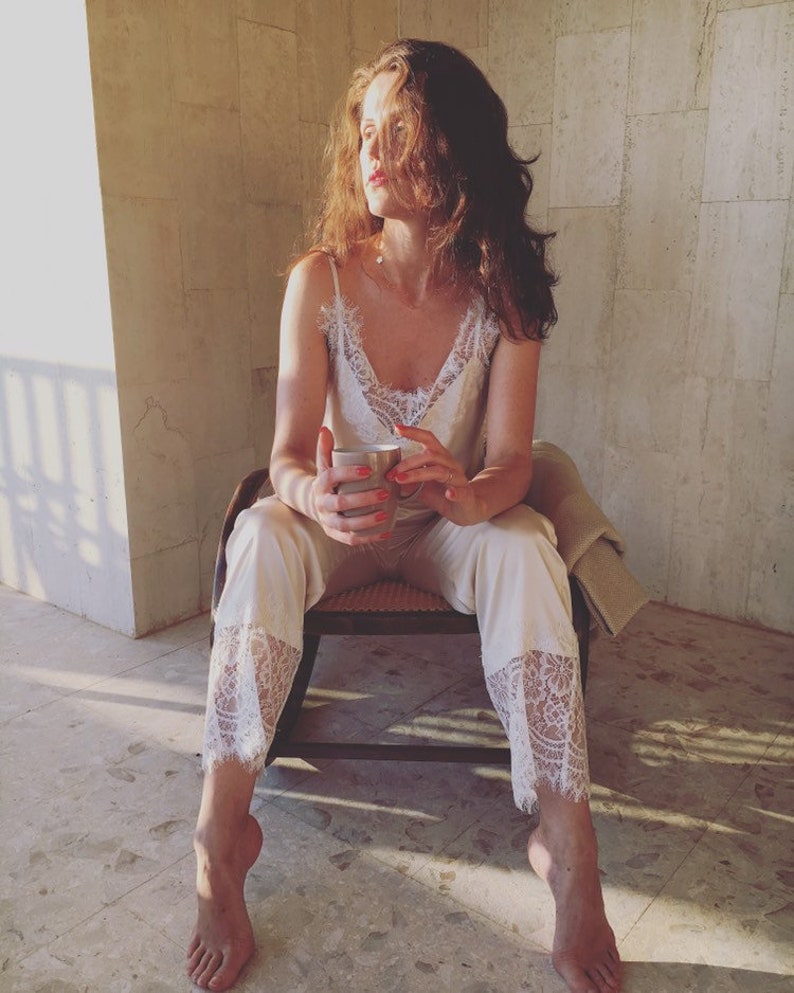Puede incluir: Una mujer con un top y pantalones de encaje blanco se sienta en una silla, sosteniendo una taza de cer&aacute;mica. La mujer mira hacia un lado y la luz le da en la cara y el pelo.