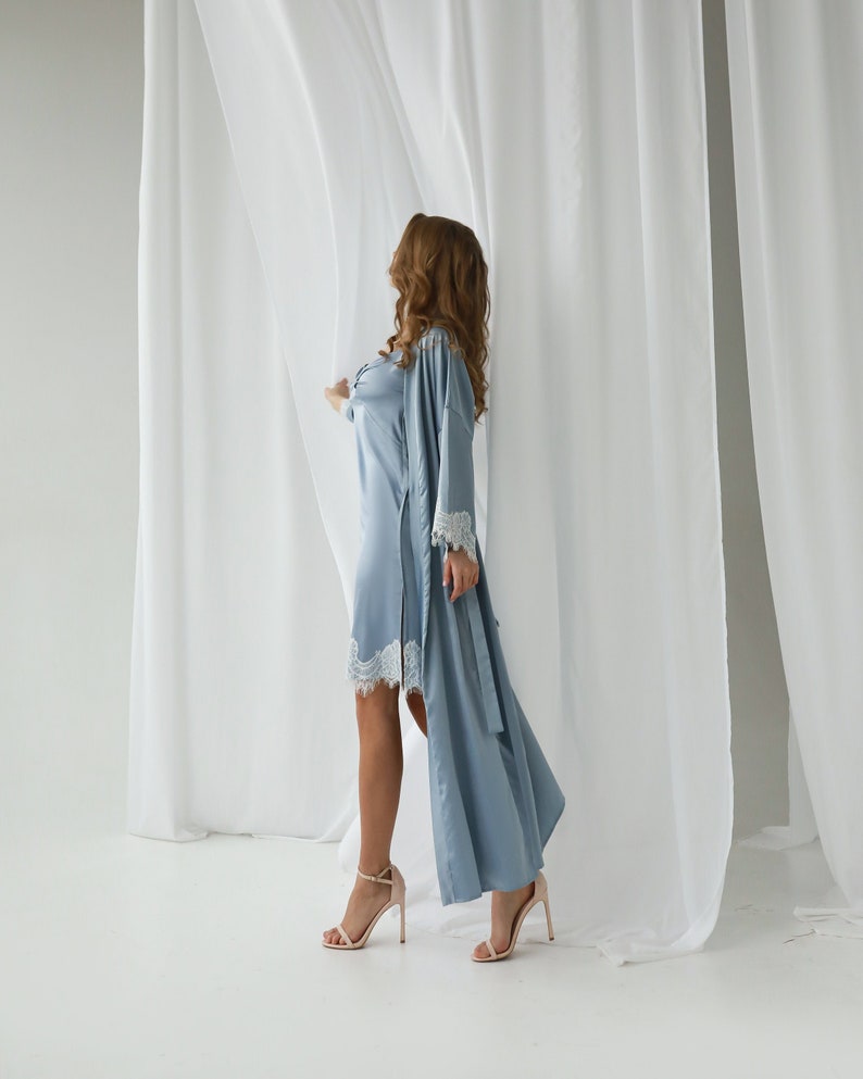 May include: A woman in a light blue satin robe with lace trim stands in front of a white curtain. She is wearing nude heels.