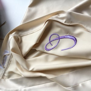 K&ouml;nnte beinhalten: Eine cremefarbene Satin-Robe mit einem gestickten, violetten Buchstaben "C" auf der Vorderseite.