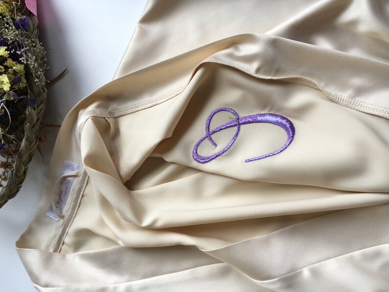 K&ouml;nnte beinhalten: Eine cremefarbene Satin-Robe mit einem gestickten, violetten Buchstaben "C" auf der Vorderseite.