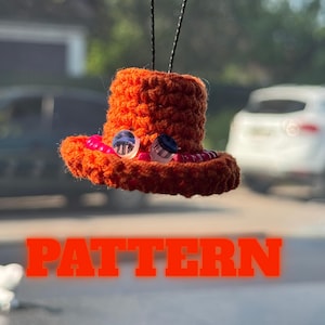 Peut inclure: Un chapeau orange au crochet avec un large bord, orné de deux perles de verre et d'une ficelle pour suspendre. Le mot "PATTERN" est affiché en grandes lettres oranges en bas de l'image.