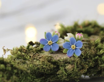 May include: A pair of blue forget-me-not flower earrings with yellow centers, displayed on a bed of green moss.
