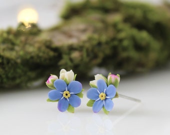 May include: A pair of delicate forget-me-not flower earrings with blue petals, yellow centers, and white and pink buds. The earrings are made of polymer clay and have silver posts.