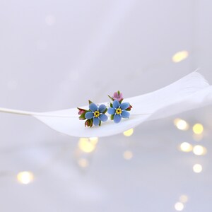 May include: A pair of blue forget-me-not flower earrings with yellow centers and green leaves. The earrings are on a white feather.