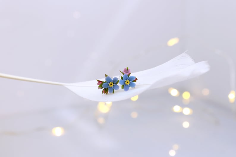 May include: A pair of blue forget-me-not flower earrings with yellow centers and green leaves. The earrings are on a white feather.