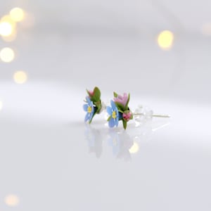 May include: A pair of delicate floral earrings featuring blue forget-me-nots and pink flowers with green leaves. The earrings are on a white surface.