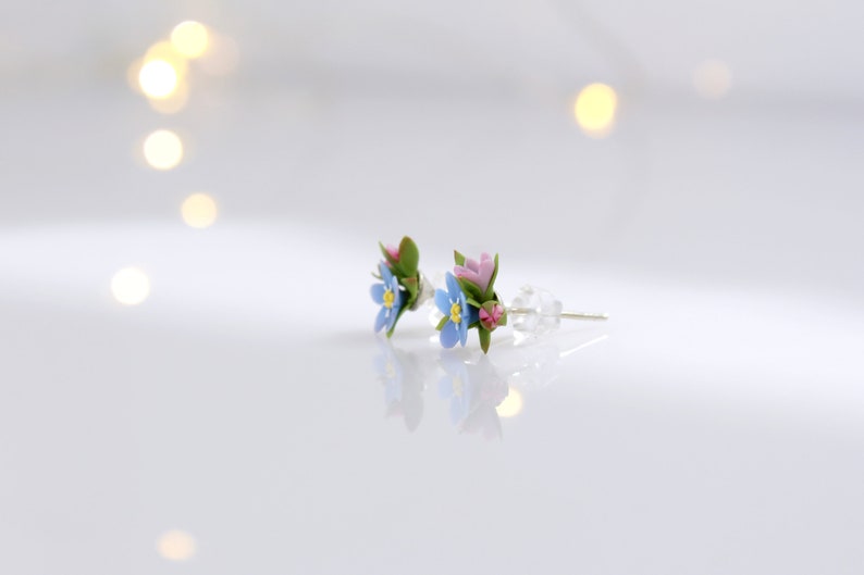 May include: A pair of delicate floral earrings featuring blue forget-me-nots and pink flowers with green leaves. The earrings are on a white surface.