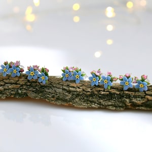 May include: Five pairs of blue and pink forget-me-not flower earrings on a piece of driftwood.