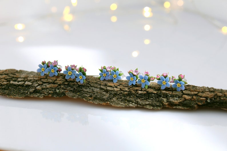 May include: Five pairs of blue and pink forget-me-not flower earrings on a piece of driftwood.
