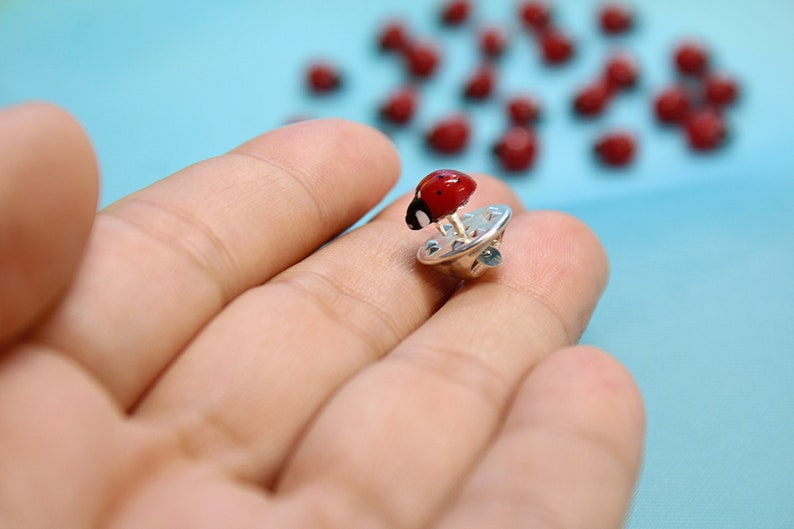 May include: A red ladybug pin with black spots, attached to a silver pin back. The pin is held in a hand against a blue background.