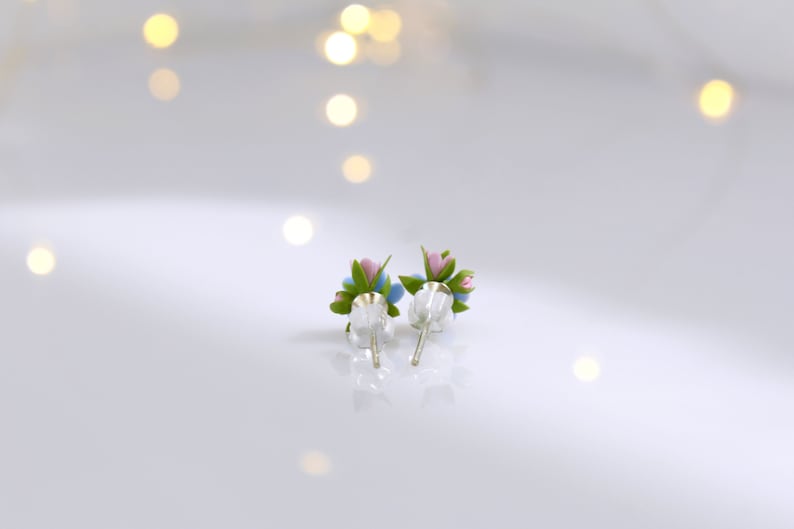 May include: A pair of silver stud earrings with small blue and pink flower accents. The flowers have green leaves.
