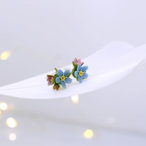 May include: A pair of delicate earrings featuring forget-me-not flowers with blue petals, yellow centers, and pink accents. The earrings are displayed on a white feather.