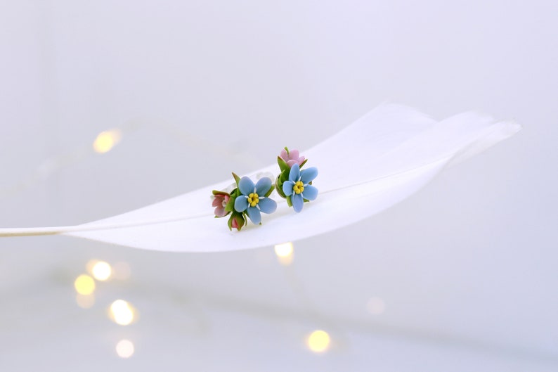 May include: A pair of delicate earrings featuring forget-me-not flowers with blue petals, yellow centers, and pink accents. The earrings are displayed on a white feather.
