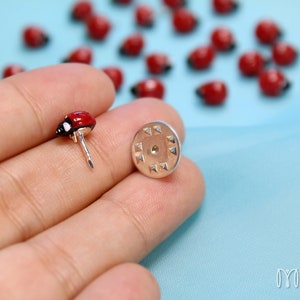 May include: A red ladybug pin with black spots and silver backing. The pin is held in a hand against a light blue background.