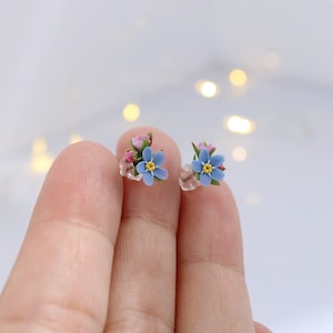 May include: A pair of delicate stud earrings featuring tiny forget-me-not flowers with blue petals, yellow centers, and pink accents.