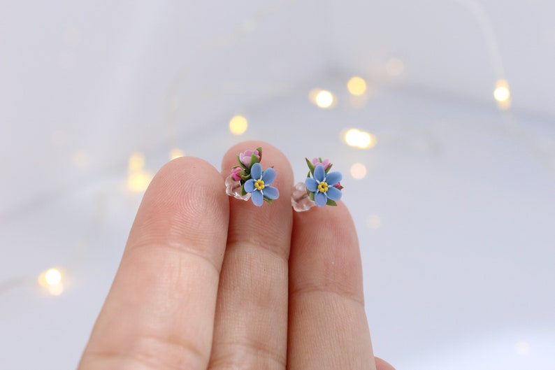May include: A pair of delicate stud earrings featuring tiny forget-me-not flowers with blue petals, yellow centers, and pink accents.