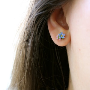 May include: A pair of small, delicate earrings featuring a blue forget-me-not flower with a yellow center and pink accents.