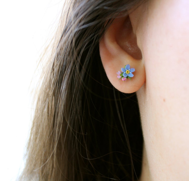 May include: A pair of small, delicate earrings featuring a blue forget-me-not flower with a yellow center and pink accents.