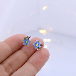 May include: A pair of delicate earrings featuring forget-me-not flowers made of polymer clay. The flowers are a light blue color with yellow centers and green stems.