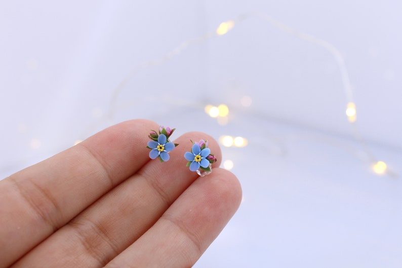 May include: A pair of delicate earrings featuring forget-me-not flowers made of polymer clay. The flowers are a light blue color with yellow centers and green stems.