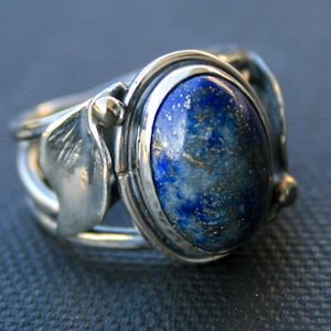 May include: A silver ring with an oval, deep blue lapis lazuli stone. The ring features leaf-like designs on the sides and small gold accents. The stone has a speckled appearance.