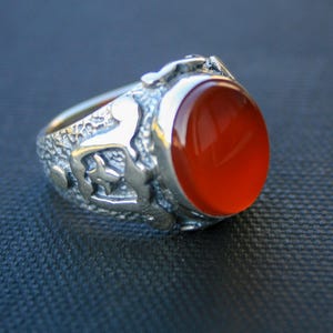 Könnte beinhalten: Ein silberner Ring mit einem ovalen, orangeroten Stein. Das Band des Rings hat ein detailliertes, strukturiertes Design mit kleinen Figuren. Der Stein ist poliert und reflektiert das Licht, vor dunklem Hintergrund. Ein Schmuckstück.