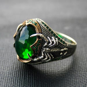 May include: A silver ring with a large, oval-cut green gemstone. The ring features a scorpion design on the band and smaller green stones around the main stone. The setting has gold accents.