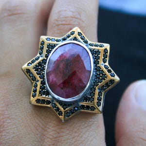 Op de afbeelding: Een goudkleurige ring met een grote, ovale, rode edelsteen in het midden. De ring heeft een stervormig ontwerp met zwarte accenten rond de edelsteen en op de sterpunten. Sieraden.