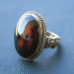 Op de afbeelding: Een zilveren ring met een ovale, donker amberkleurige steen. De steen is gezet in een getextureerde, touwachtige bezel. De band heeft Keltische knoopontwerpen. De ring ligt op een blauw oppervlak.