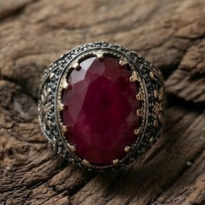 May include: A silver ring featuring a large, oval-shaped, faceted deep red gemstone set in a gold-toned bezel. The band is crafted with intricate, dark oxidized silver metalwork, displaying detailed floral engravings and a textured, granular finish.