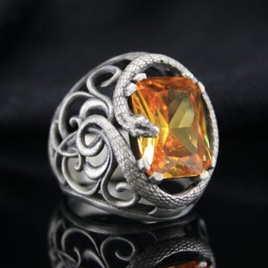 Può includere: Un anello color argento con un serpente testurizzato e intricato avvolto attorno a una grande pietra preziosa arancione rettangolare sfaccettata. La fascia presenta un design traforato con motivi a volute simili a viticci.