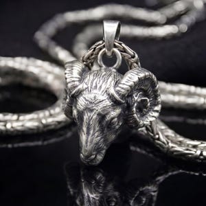 Op de afbeelding: Een zilverkleurige hangerketting met een gedetailleerde rammenkop. De rammenkophanger is bevestigd aan een dikke, getextureerde ketting. De donkere achtergrond benadrukt de metalen glans van de ketting. De ketting is een sieraad.